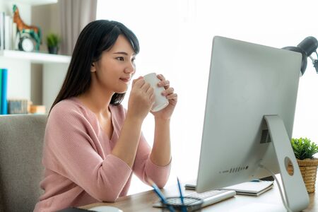 Asian beautiful young woman work from home working on computer and drinking coffee while working in living room at home. Social distancing lifestyle in house.の写真素材