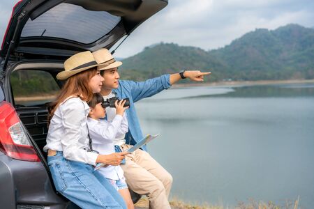 Portrait of Asian family sitting in car with father pointing to view and mother holding maps with daughter looking beautiful landscape through binoculars while vacation together in holiday. Happy family time.の写真素材