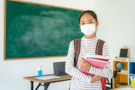Asian primary students girl with backpack and books wearing masks to prevent the outbreak of Covid 19 in classroom while back to school reopen their school, New normal for education concept.の写真素材