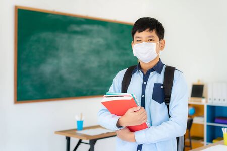 Asian primary students boy with backpack and books wearing masks to prevent the outbreak of Covid 19 in classroom while back to school reopen their school, New normal for education concept.の写真素材