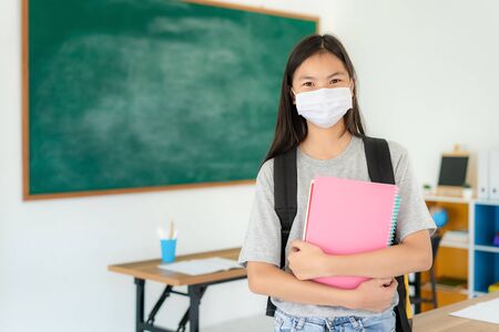 Asian primary students girl with backpack and books wearing masks to prevent the outbreak of Covid 19 in classroom while back to school reopen their school, New normal for education concept.の写真素材