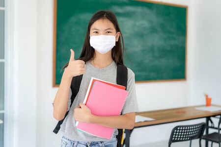 Asian primary students girl thumb up and  wearing masks to prevent the outbreak of Covid 19 in classroom while back to school reopen their school, New normal for education concept.の写真素材