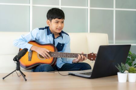 Asian boy playing acoustic guitar and watching online course on laptop while practicing at home. Asian boy virtual happy hour meeting for play music online together with friend via videocall.の写真素材
