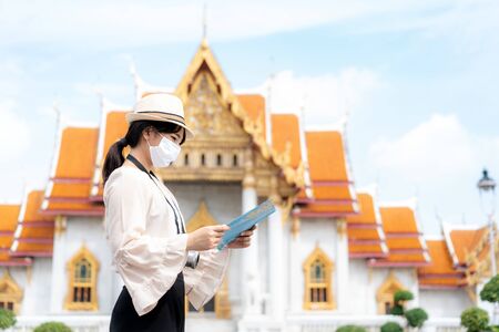 Asian woman happy tourists to travel wearing mask to protect from Covid-19 on her holidays and she looking at travel map in Temple in Bangkok, Thailandの写真素材