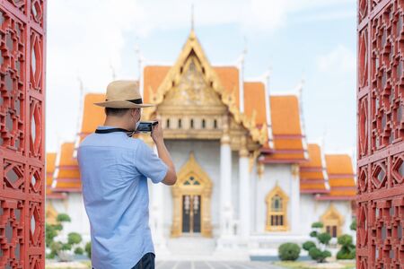 Asian man happy tourists to travel wearing mask to protect from Covid-19 on his holidays and he taking a photo by camera in Temple in Bangkok, Thailandの写真素材