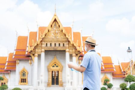 Asian man happy tourists to travel wearing mask to protect from Covid-19 on his holidays and he looking at travel map in Temple in Bangkok, Thailandの写真素材