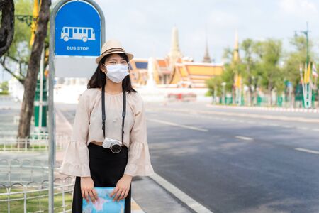 Asian woman happy tourists to travel wearing mask to protect from Covid-19 on her holidays and she looking bus at bus stop in Wat Phra Kaew Temple in Bangkok, Thailandの写真素材