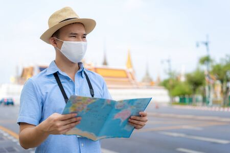 Asian man happy tourists to travel wearing mask to protect from Covid-19 on his holidays and he looking at travel map in Wat Phra Kaew Temple in Bangkok, Thailandの写真素材