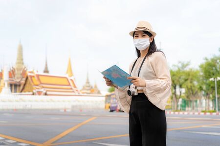 Asian woman happy tourists to travel wearing mask to protect from Covid-19 on her holidays and she looking at travel map in Wat Phra Kaew Temple in Bangkok, Thailandの写真素材