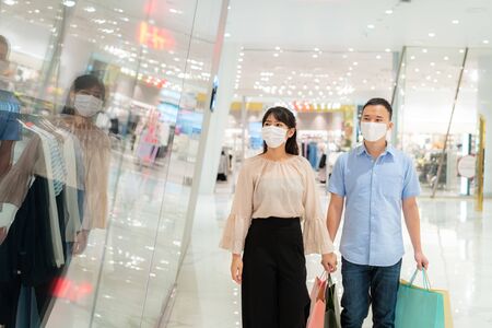 Asian couple wearing mask over their face looking new collection dress at shopping mall with shopping bag for healthcare and prevention from coronavirus, Covid19 influenza in crowded place.の写真素材