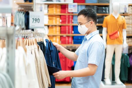 Asian man wearing mask over her face while choosing shirt at shopping mall with shopping bag for healthcare and prevention from coronavirus, Covid19 influenza in crowded place.の写真素材