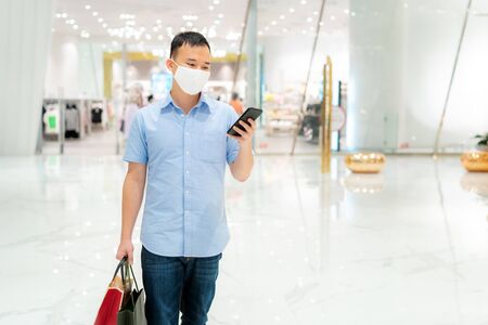 Asian man wearing mask over his face while walking at shopping mall with shopping bag for healthcare and prevention from coronavirus, Covid19 influenza in crowded place.の写真素材