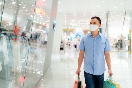 Asian man wearing mask over his face while walking at shopping mall with shopping bag for healthcare and prevention from coronavirus, Covid19 influenza in crowded place.の写真素材