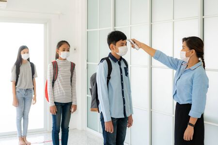 Asian woman teacher using thermometer temperature screening student for fever against the spread of COVID-19 while student coming back to school, New normal and Education concept.の写真素材