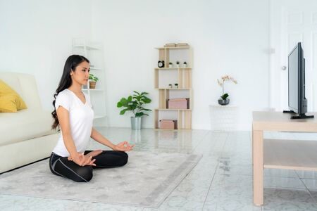 Beautiful young Asian woman in sports clothing doing yoga while relaxing in living room at home to get exercise into her daily routine while social distancing.の写真素材