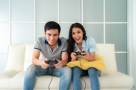 Asian couple man ans woman to playing video games with joysticks while sitting in sofa in living room at home, concept of family values and the modern-day game.の写真素材
