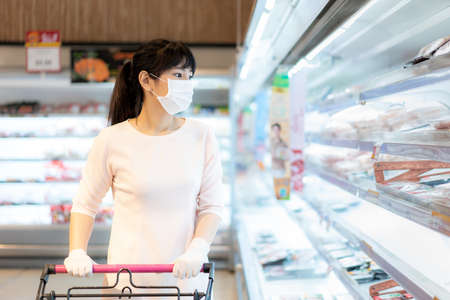 Asian woman with hygienic mask and rubber glove with shopping cart in grocery and looking for fresh meat pack to buy during covid-19 outbreak for preparation for a pandemic quarantineの写真素材