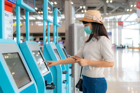 Asian woman traverler wearing mask using self check-in kiosk in airport terminal during coronavirus (COVID-19) pandemic prevention when travel abroad. New normal travel  conceptの写真素材