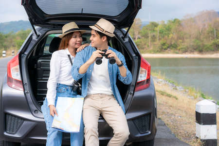 Young Asian couple with man tourist holding binoculars with smiling girlfriend sitting near with map...
