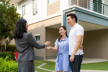 Asian happy smile young couple take keys new big house from real estate agent or realtor in front of their house after signing contract agreement, concept for real estate, moving home or renting propertyの写真素材