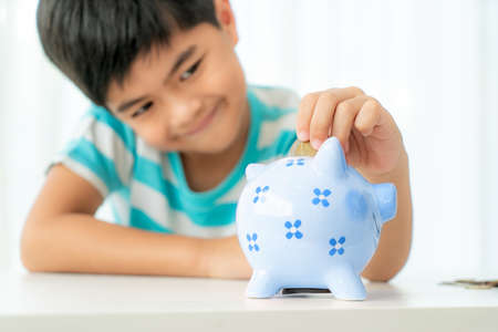 Little Asian boy insert a coin into blue piggy bank in white table at living room at home for the kid saving, investment money for his business future conceptの写真素材