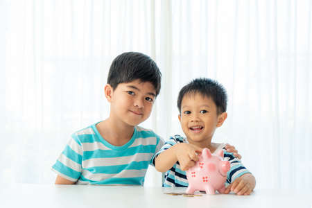 Two Asian cute brother and son insert coin  in piggy bank together on the table in living room at home. Concept of bonding of sibling, friendship and saving, investment money for his business future conceptの写真素材