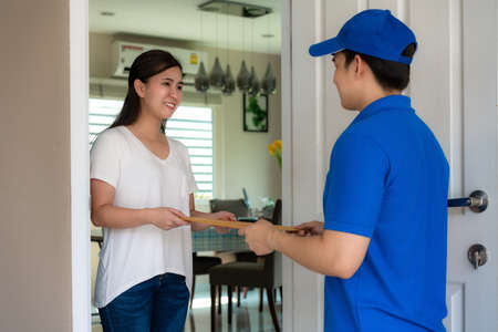 Asian delivery young man in blue uniform smile and holding letter or document envelope in front house and Asian woman accepting a delivery of envelope from deliveryman. Advertising, Transportation.の写真素材