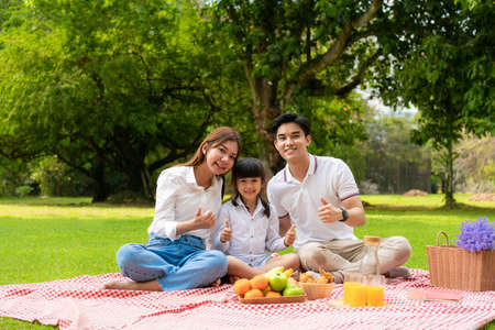 Asian teen family happy holiday picnic moment in the park with father, mother and daughter looking at camera and smile to happy spend vacation time togerter in green garden with fruit and food.の写真素材