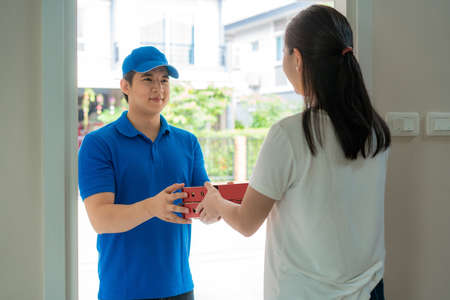 Asian delivery young man in blue uniform smile and holding pizza boxes in front house and Asian woman accepting a delivery of pizza boxes from deliveryman. Advertising, Business, Transportation.の写真素材