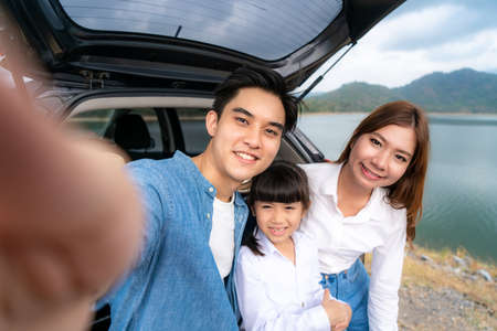 Portrait of Asian family sitting in car with father, mother and daughter selfie with lake and mountain view by smrtphone while vacation together in holiday. Happy family time.の写真素材