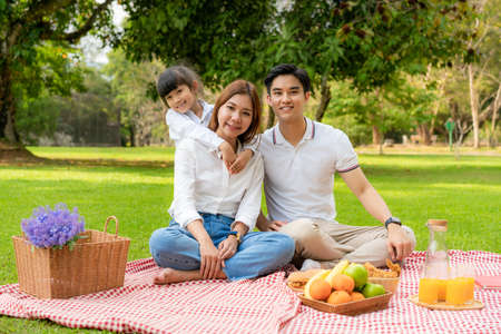 Asian teen family happy holiday picnic moment in the park with father, mother and daughter looking at camera and smile to happy spend vacation time togerter in green garden with fruit and food.の写真素材