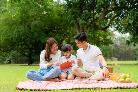 Asian teen family happy holiday picnic moment in the park with father, mother and daughter reading book togerter in green garden with fruit and food.の写真素材