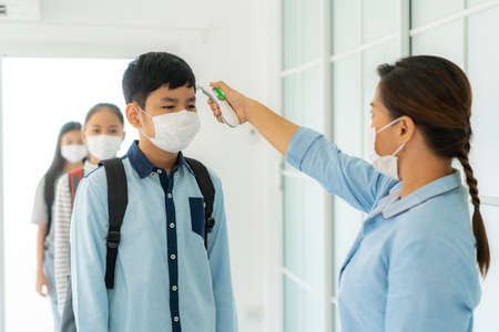 Asian woman teacher using thermometer temperature screening student for fever against the spread of COVID-19 while student coming back to school, New normal and Education concept.の写真素材