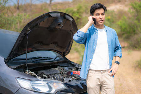 Asian young unhappy man talking on a cell phone  in front of the open hood  broken down car On Country Road Phoning For Help. Broken Car On The Road in During journey.の写真素材
