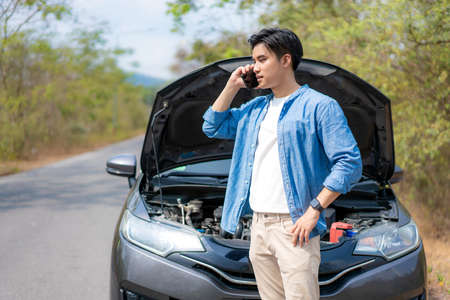 Asian young unhappy man talking on a cell phone  in front of the open hood  broken down car On Country Road Phoning For Help. Broken Car On The Road in During journey.の写真素材