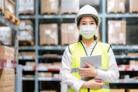 Young asian woman auditor or trainee staff wears mask working during the COVID pandemic in store warehouse shipping industrial. looking up and checks the number of items store by digital tablet.の写真素材