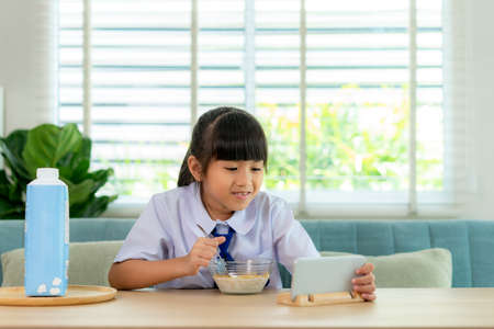 Asian elementary school student girl in uniform eating breakfast cereals with milk and looking cartoon in smartphone to distractions to eat it in morning school routine for day in life getting ready for school.の写真素材