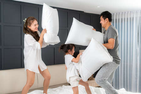 Asian happy family having funny pillow fight on white bed in bedroom for free time with father, mother and daughters feeling happy and family relationship in bedroom at home.の写真素材