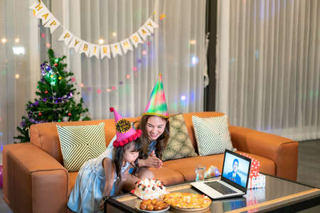 Happy little girl celebrating birthday with her mother at home with father on video call in laptop while blowing candles in cake on table.の写真素材
