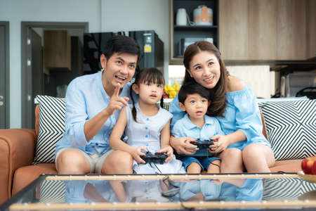 Happy asian family with father, mother, daughter and son playing games using joystick having funny moment together on sofa in living room at home.の写真素材