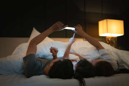 Asian happy family mother, father and little daughter watching movie or cartoon in smartphone together and blanket cover their head in bed at night at homeの写真素材