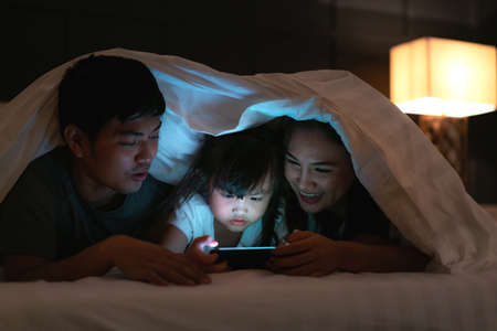 Asian happy family mother, father and little daughter watching movie or cartoon in smartphone...