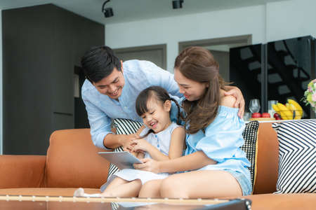 Happy Asian family with father, mother and daughter sitting on sofa and using digital tablet looking at screen together in living room at home.の写真素材