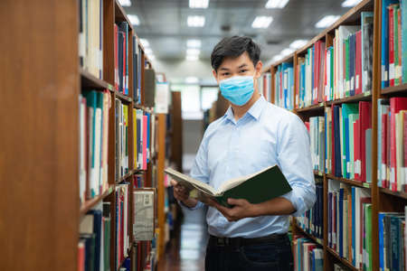 Asian university student boy wearing face protective medical mask for protection from virus disease reading book at library. Education, high school, university, learning and people conceptの写真素材