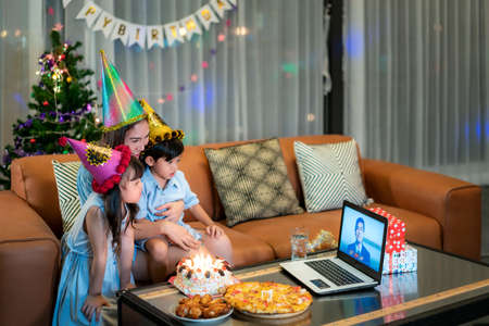 Happy little girl and son celebrating birthday with her mother at home with father on video call in laptop while blowing candles in cake on table.の写真素材