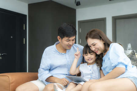 Happy Asian family with father, mother and daughter sitting on sofa and using digital tablet looking at screen together in living room at home.の写真素材