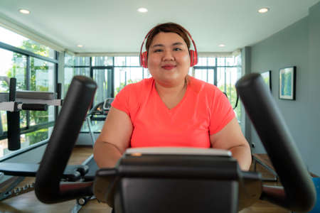 Happy Asian Overweight woman with earphone training on exercise bike in modern gym, happy and smile during workout. Fat woman take care of health and want to lose weight concept.の写真素材