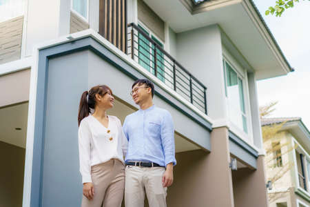 Portrait of Asian young couple standing and hugging together looking happy in front of their new house to start new life. Family, age, home, real estate and people concept.の写真素材