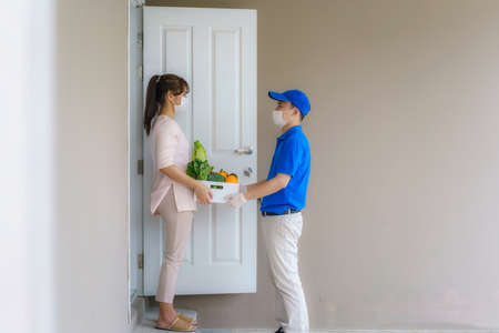 Asian woman costumer wearing face mask and glove receive groceries box of food, fruit, vegetable and drink from delivery man   in front of the house during time of home isolation.の写真素材