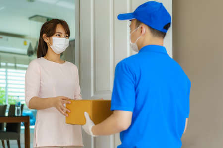 Asian delivery man wearing face mask and glove in blue uniform holding a cardboard boxes in front house and woman accepting a delivery of boxes from deliveryman during COVID-19 outbreak.の写真素材